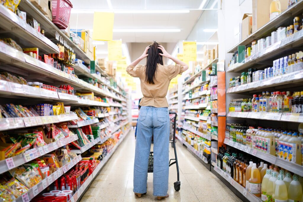 A woman in a Japanese supermarket holds her head in frustration while looking at grocery shelves, symbolizing rising prices and inflation concerns.